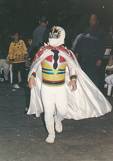 a dwarf dressed in a white body suit and mask, wearing a colorful cape on the way to the ring.