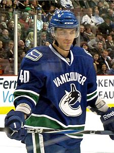 A Caucasian male ice hockey player in his mid-twenties. He stands relaxed on the ice, holding his hockey stick horizontally across his hips. He wears a blue, visored helmet and a blue jersey with green and white trim.