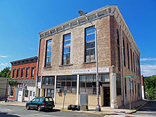 Two brick buildings seen from across the street and slightly to their left. The one closer to the camera has a higher second story with long tall windows. Its bricks have traces of a past white covering.