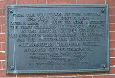 An image of darkened brass historical plaque with a streak of green corrosion running down it, mounted on the exterior side of a brick building.
