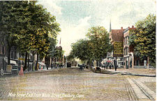 A hand-tinted postcard image showing a cobblestone-paved, tree-lined street with many commercial buildings and some horse-drawn carriages. White script at the bottom left corner reads "Main Street, East from White Street, Danbury, Conn."