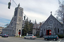 Two pointed gray stone buildings seen from across a street, with cars passing in front and a traffic light visible from the center to the upper left. The one on the left has a tall square stone tower with a smaller tower topped by a cross at the top. The one on the left has a large round window in the middle and a cross at the point on the front. Both have open red doors.