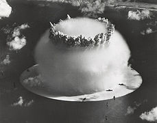 Aerial photo of nuclear explosion rising from lagoon. Hemispherical condensation cloud on the surface is 1 mile (1.6 km) in diameter. In comparison, Navy ships in the foreground look like bathtub toys.