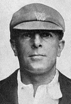 Portrait of man from just below the shoulder upwards. He is wearing a light shirt and blazer over the top, a cricket cap, dark with a white crest. The resolution is poor and nothing more of the crest can be discerned. He is unshaven and staring at the camera, not smiling.
