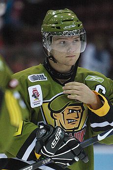 A frontal view of a white, teenage ice hockey player during a game. He is wearing a green, visored helmet and an olive green jersey with a logo consisting of an animated face of a soldier. He appears calm while looking to the right and is reaching for his face with his right hand.