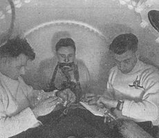 Three men inside a pressure chamber. One is breathing from a mask and the other two are timing and taking notes.