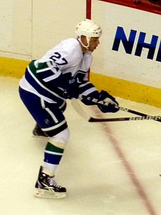 An ice hockey player seen from the side in a ready position. His legs are planted as he holds his stick outward in front of him. He wears a white jersey with blue and green trim.