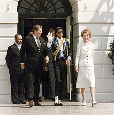 In the center of the photo four people can be seen. To the farthest left a medium skin colored man wearing a black suit with a white shirt can be seen. Second to the left a Caucasian man wearing a black suit with a white shirt and brown tie has his head turned to the right. To the right of the Caucasian male there is an African American man wearing a white shirt with a blue jacket that has a yellow strap across his chest. He is raising his right hand, which is covered with a white glove. To the farthest right, a Caucasian female with short blonde hair, who is wearing a white outfit, can be seen. In the background a cream colored building with an opened green door can be seen.