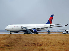 A large white jet with a red-and-blue tail on a runway amid a yellowed field of grass. A gray truck has extended a ladder to the plane's door.