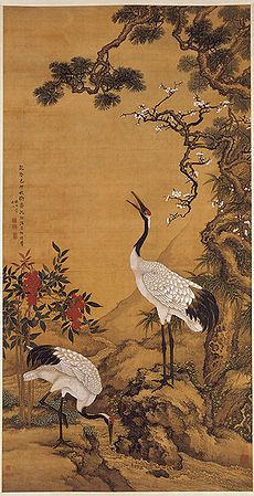 Two cranes near a pine tree. One is feeding on the ground while another rears its head high. Red flowers are also in the background