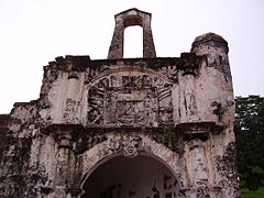 Stained ruin of a stone building, showing a central arch, flanked by two columns, with a stone relief above the arch, also flanked by two columns, and a second free-standing arch perched on the very top of the ruin.