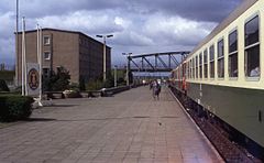 View of a train stopped at a long railway platform, at the end of which is an arched iron bridge. A grey concrete barracks and East German state emblem are visible on the side of the platform. Several people are standing or walking on the platform and the train's doors stand open.
