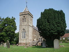 Gray stone building. Prominent square tower with arched window, topped by small slate pyramidal roof. Left and right of the building are yew trees amongst gravestones.