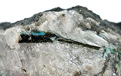 Photograph of a mendipite sample with a dark crystal of chloroxiphite embedded in it and a smaller, bright blue diaboleite crystal at the chloroxiphite's top edge.