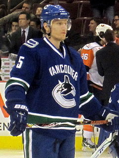 A Caucasian ice hockey player in his late-twenties looking forwards.  He is wearing a blue jersey with green and white trim, as well as a blue, visored helmet.  He holds his hockey stick horizontally across his waist in a relaxed position.