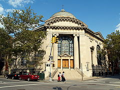 The front entrance of a hexagonal building capped by a dome is visible, facing a street-corner. The entranceway is framed by large stone columns and flanked by metal seven branched menorahs on each side. There are four wooden doors, one on each side and two in the middle, topped by a large arched stained-glass window. A stone stairway with metal railings on each side leads up from the sidewalk to the doors.