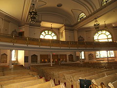 The back of a wide, two to three-story room is visible. Four visible rows of wooden pews lead to the back wall, which has three sets of double doors at its center. A second-floor balcony which projects partway into the sanctuary holds more wooden pews. The back wall has two large arched stained-glass windows visible, and two smaller rectangular ones one each side of the doors. The ceiling is arched, with elaborate chandeliers with Star-of-David shapes hanging from it.