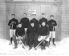 Seven men in hockey uniforms are arranged in two rows in front of a large door, on which are the words "Dey's Rink" In the middle of the front row, is their manager, wearing a dark coat and bowler hat.