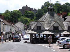 Street scene with houses and shops on the left and an octagonal structure has a central stone pier which supports a heavy timber framework which carries a slate roof with central wooden lantern surmounted by a weather vane. In the distance is a castle.