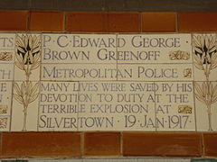 A tablet formed of five tiles of varying sizes, bordered by yellow wheatsheaves in an art nouveau style. The tablet reads "P.C. Edward George Brown Greenoff, Metropolitan Police. Many lives were saved by his devotion to duty in the terrible explosion at Silvertown 19 Jan 1917".
