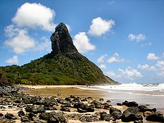 In the foreground, a wavy sea at the right and a yellow beach covered with black stones at the left. In the background, a mountain covered with green vegetation out of which towers a steep black rock.