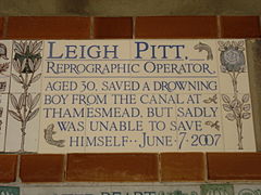Almost identical to the plaque to Amelia Kennedy, a tablet formed of five tiles of varying sizes, bordered by yellow and blue flowers in an art nouveau style and decorated by two stylised salmon. The tablet reads "Leigh Pitt, reprographic operator, aged 30, saved a drowning boy from the canal at Thamesmead, but sadly was unable to save himself June 7, 2007".