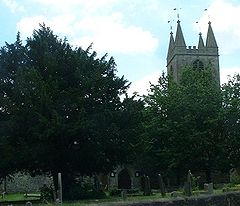 Top of ower with spirelets seen behind trees. In the foreground is grass and gravestones