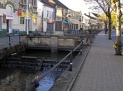 A river running between pavements with railings. Shops behind