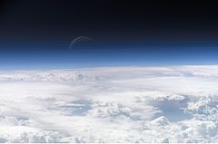 White cloud tops below with Earth's azure atmosphere blending into outer space, where a thin crescent moon hovers against the darkness
