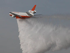 A three-engine red-and-white cargo plane in-flight, releasing a large quantity of water from its undercarriage storage tanks. The water trails behind the aircraft in a continuous, fan-shaped drop pattern.