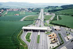 Aerial view of a four-lane motorway crossing green fields, with a small village with a church spire in the distance to the left of the motorway. In the foreground, there is a white roof structure, resting on slim white pillars, across all four lanes of the motorway; to the left, the roof also extends over a slip lane which branches off from the main road and then rejoins it; on the right, just before the roof structure, there is a parking lot with diagonally parked orange and brown lorries.