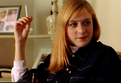 Photo of a young strawberry-blonde woman with a smile on her face; she is clad in a dark jean jacket and holding a cigarette.