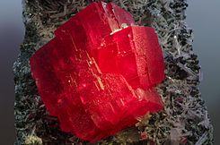 alt1=A mass of blocky, vivid red crystal extends from a dark rock covered with small, translucent white, rodlike crystals