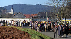 A large number of people of various ages standing and walking along a road in front of a high concrete wall, behind which houses and a church are visible in a wooded valley.