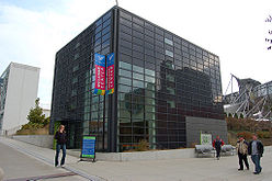 A cube-shaped building covered by a rectangular grid containing some windows, but mostly black glass. Four people are on the surrounding sidewalks and there are three banners reading "Welcome Center".