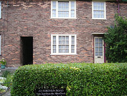 Exterior of a two-story brick building, with a hedge in front of it. Six windows are visible, three on each level, as are two doorways on the lower level.