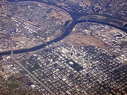High angled aerial shot of a developed city; a suburban grid dominates the lower half of the image. A river bisects the city from the left before forking; the first fork continues up and to the right edge of the image; the second curves up and around to finish on the left, enclosing industrial units and other domestic properties.