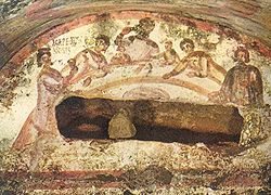 Mural painting in a Roman catacomb showing five adults and two children at a table. One of the adults holds up a chalice. The painting is damaged by the opening of the wall to remove the remains that were interred behind the picture.