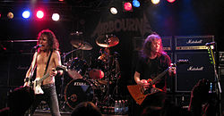 Three members of the band are in view. The man at left is singing into a microphone while playing his guitar. He is bare-chested, has shoulder length curling dark hair and wears dark pants. The second man is obscured behind his drum kit at mid-stage. The man at right is playing his guitar. He has his eyes partly closed, has over shoulder length hair and is slightly turned to his left. He wears a dark tee shirt and dark pants. Behind the musicians are large speakers and stage lights of white, red and blue are overhead. The band's name 'Airbourne' is displayed on a back-board, above the drummer.