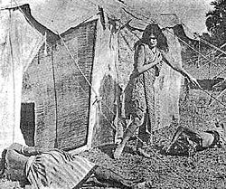 A young woman with long dark hair walks outside of a tent, looking down at one of two men asleep on the ground. She wears only a shawl and a knee length dress, leaving her arms, lower legs, and feet exposed.