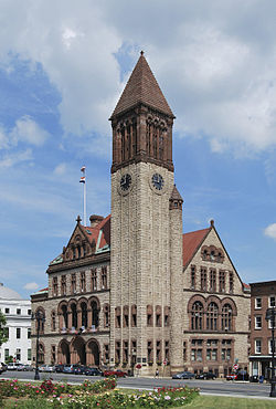 A light brown building with dark brown trim stands on a street corner; it has an arched entrance at left, a double-peaked roof, and a 200-foot tower at the closest corner.