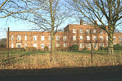 A tall wire fence separates the viewer from a long red brick multi-storey building, set in parkland.
