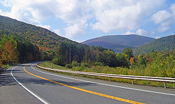A paved highway with double-yellow centerline, shoulders and a guardrail on the far side curves across the foreground. In the distance is a high, flat-topped mountain covered in woods, with some trees showing early autumn color, framed by similar slopes on either side. Above the landscape is a blue sky with clouds.