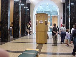 A large hall flanked by black marble columns on the left and right. There is a wooden tower in the middle on which are mounted several plaques. Two people are standing at the tower; one, obscured behind the other, points at a plaque while other people mill about on the sides of the room. The far end of the room opens into a lighted rotunda.