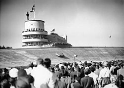 A large crowd of people watch a streamlined single-seater drive around a massive curved racing bank. A Nazi flag is flying in the distance atop a tower.
