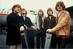 A photograph of five young men with moptop haircuts, looking windswept and standing in front of a passenger airplane. The five are all dressed in casual jackets and jeans, and three of them are resting their hands on guitar cases.