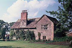 A stone house seen from a slight angle, with a gabled wing to the right and a tall chimney on the roof to the lefy