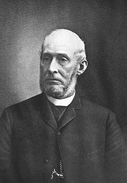 Formal portrait, head and shoulders, of a serious-looking man of about 60 dressed in a dark suit jacket and white collar. Nearly bald, he has a neatly trimmed, short gray beard.