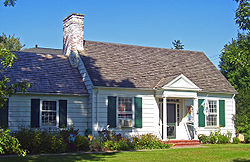 A small half-shaded white house-like building with green shutters and a white brick chimney