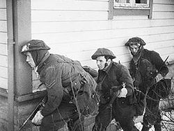 A black and white photograph of three British soldiers advancing beside a wooden house. The lead soldier is carrying a Thompson submachine gun and is peering around the corner of the house, while the other two are carrying rifles.